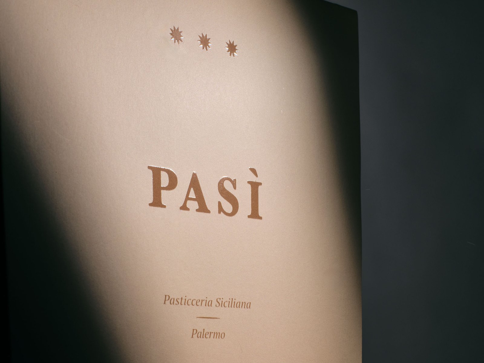 The Pasì brand mark and its three stars crossed by a blade of raking light, on a powder pink box