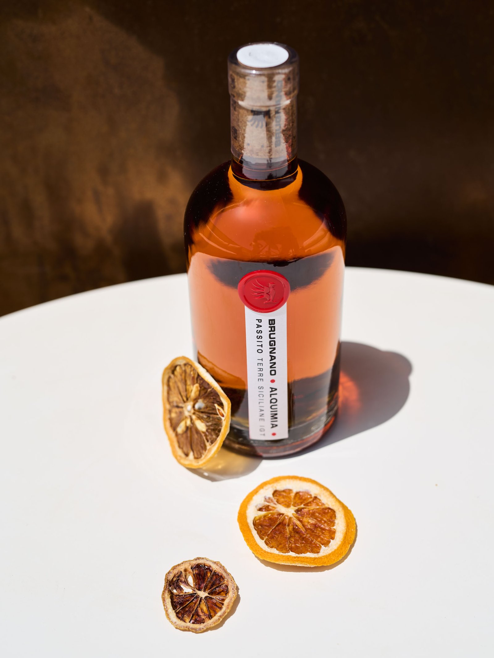 Alquimia Passito and slices of dried citrus on a white table, overhead sun