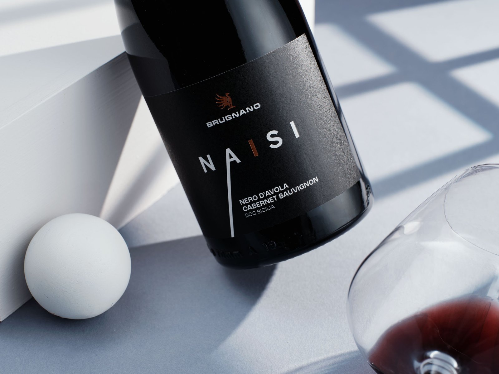The Naisi bottle on a pale grey ground, the hard shadow of a window and a glass of red