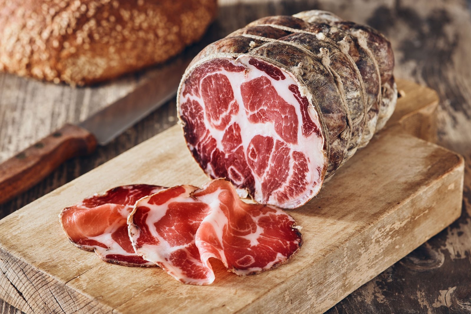 Salume stagionato legato con lo spago, aperto sul tagliere con le fette in primo piano, il pane e il coltello