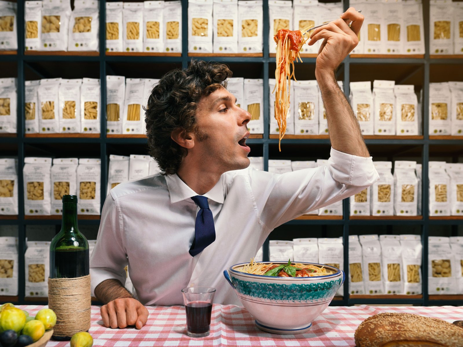 Un uomo in camicia e cravatta alza una forchettata di spaghetti al sugo di pomodoro davanti a una parete di scaffali pieni di pasta, su una tovaglia a quadri