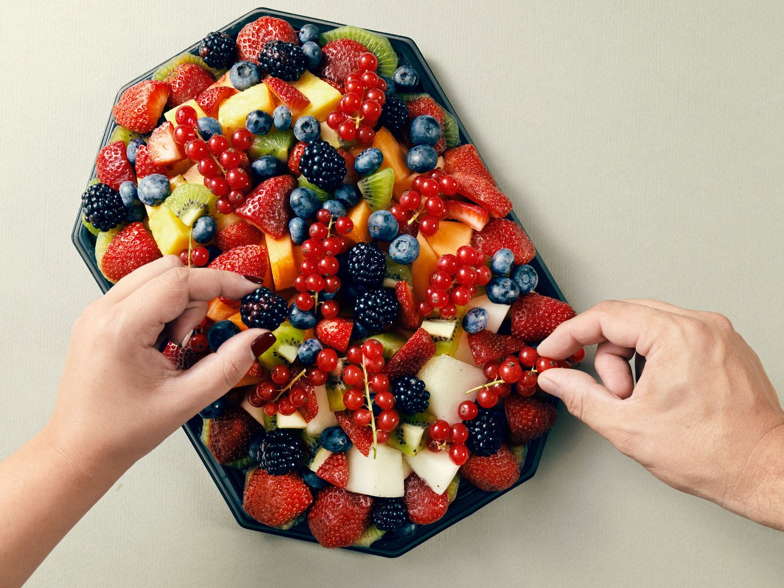 Vassoio di frutta tagliata visto dall'alto, con fragole, mirtilli, more, ribes, kiwi e melone, e due mani che ci si allungano insieme