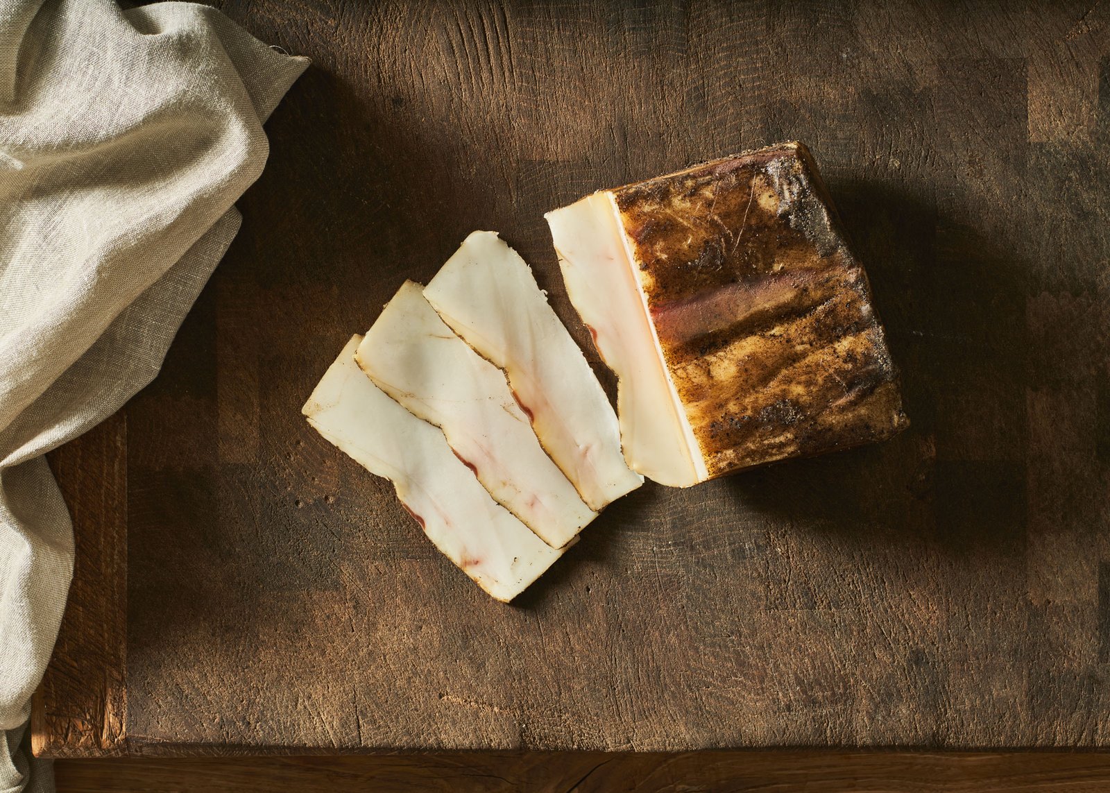 Fette di lardo e il pezzo intero con la crosta scura di spezie, luce radente sul legno