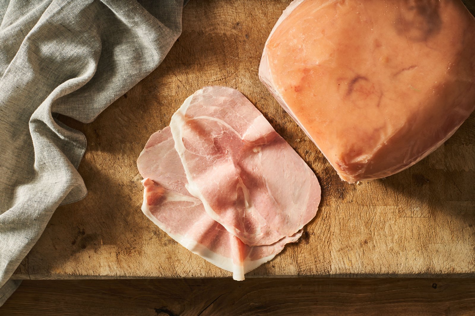 Fette di prosciutto cotto piegate accanto al pezzo intero, su tagliere e telo di lino