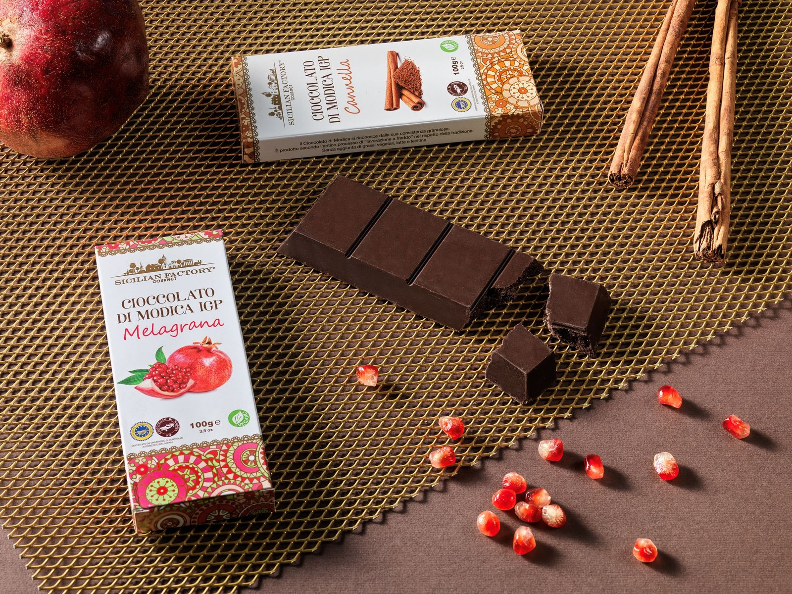 Modica PGI chocolate with pomegranate and with cinnamon: the two boxes and a broken bar on a gold mesh, with pomegranate seeds and cinnamon sticks