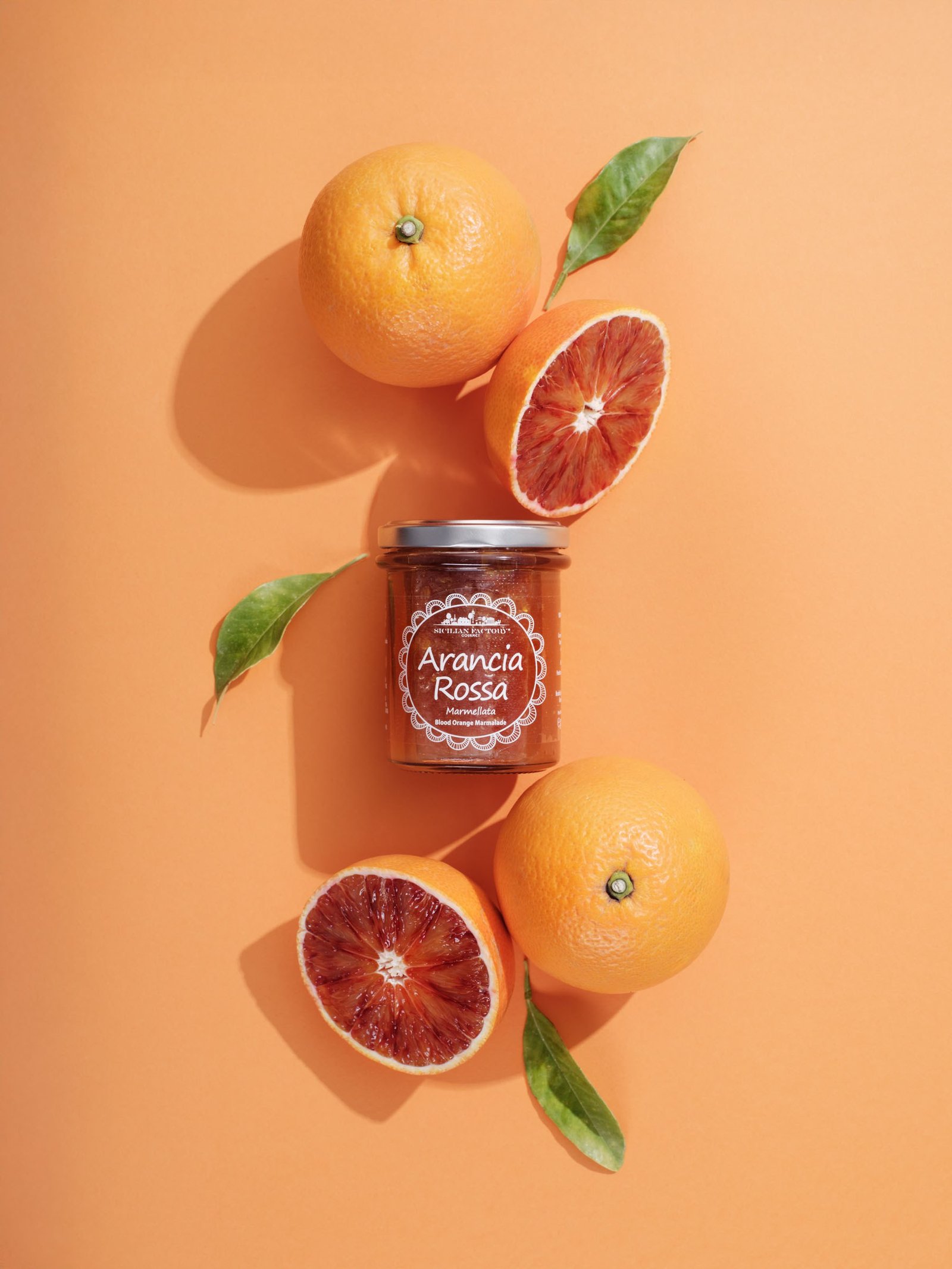 Blood orange marmalade shot from above among whole oranges, halved blood oranges and leaves, on an orange background in hard light