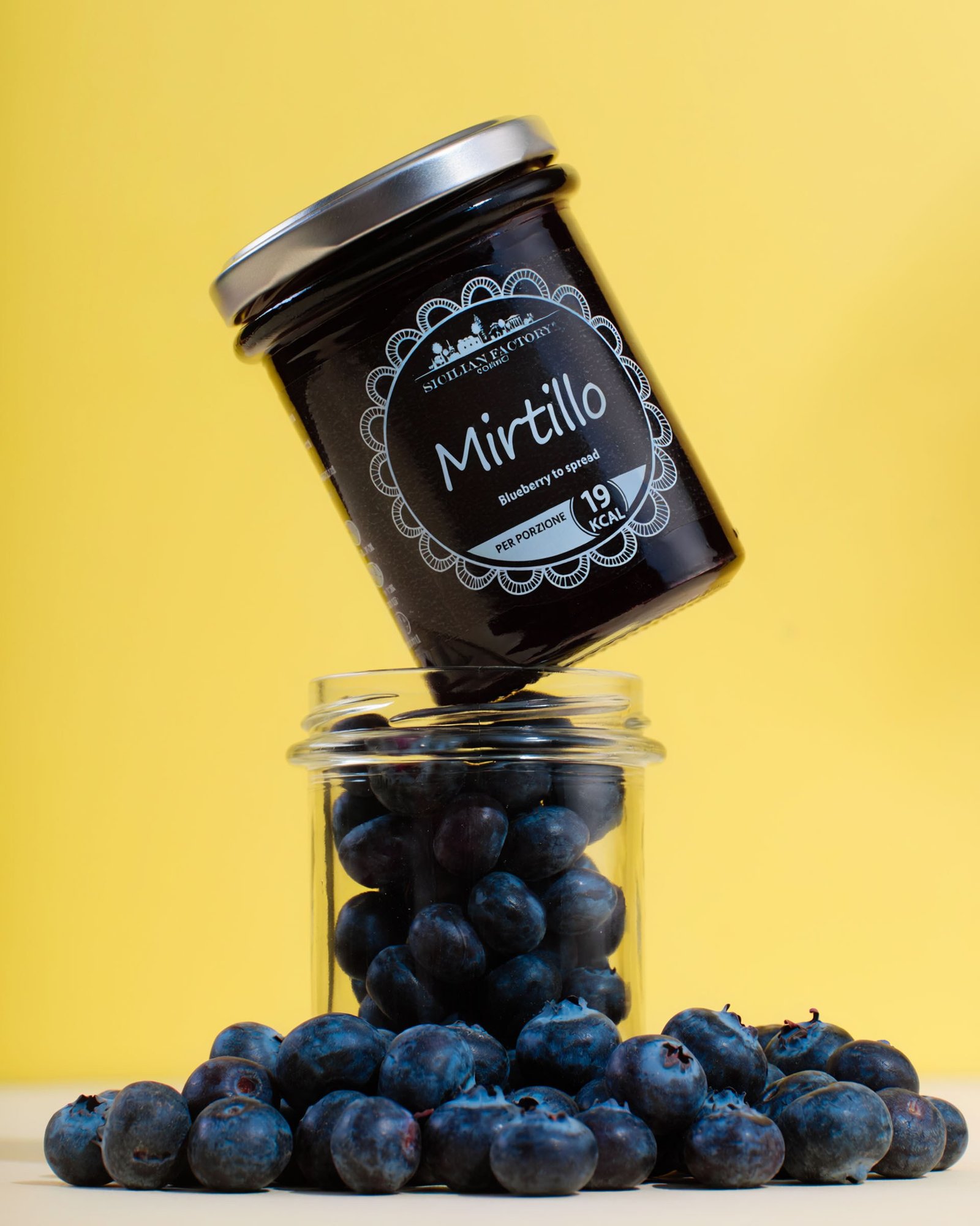 Sicilian Factory blueberry spread balanced on a glass jar full of blueberries, on a yellow background