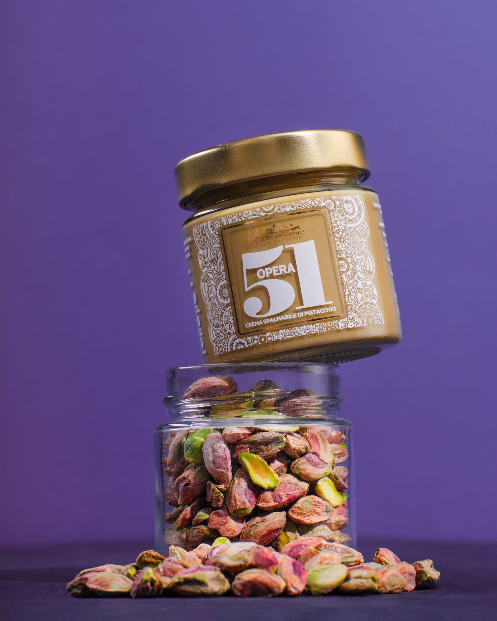 Opera 51 pistachio spread balanced on a glass jar full of pistachios, on a violet background