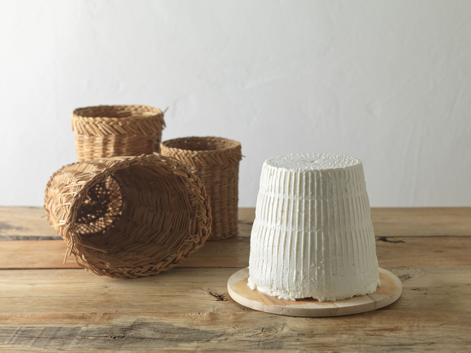 Una ricotta intera su un tagliere tondo di legno, accanto a tre cesti intrecciati, uno coricato su un fianco