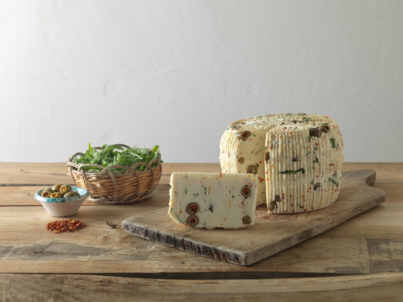 Primo sale con olive, peperoncino e rucola: nello spicchio tagliato si vedono le olive verdi; accanto la rucola in un cesto, le olive in una ciotola e il peperoncino