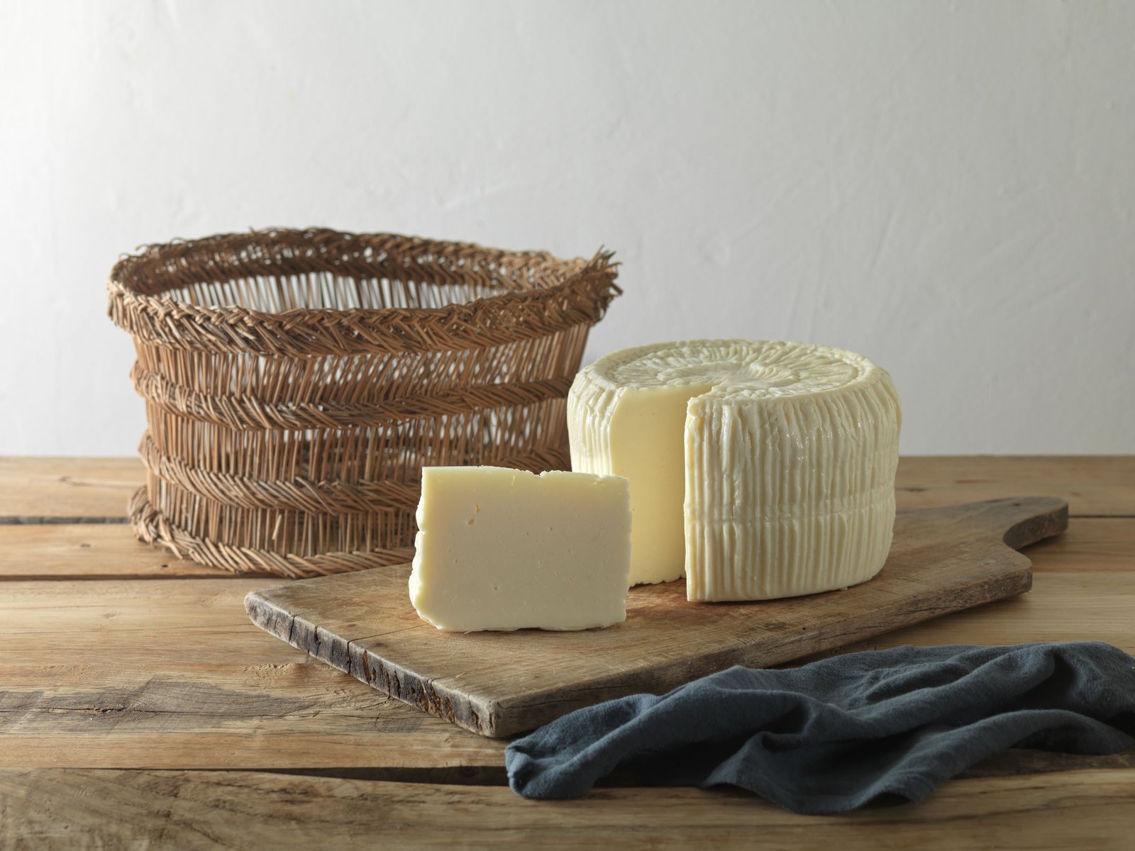 Primo sale con uno spicchio tagliato, su un tagliere di legno accanto a un grande cesto intrecciato e a un telo grigio-azzurro