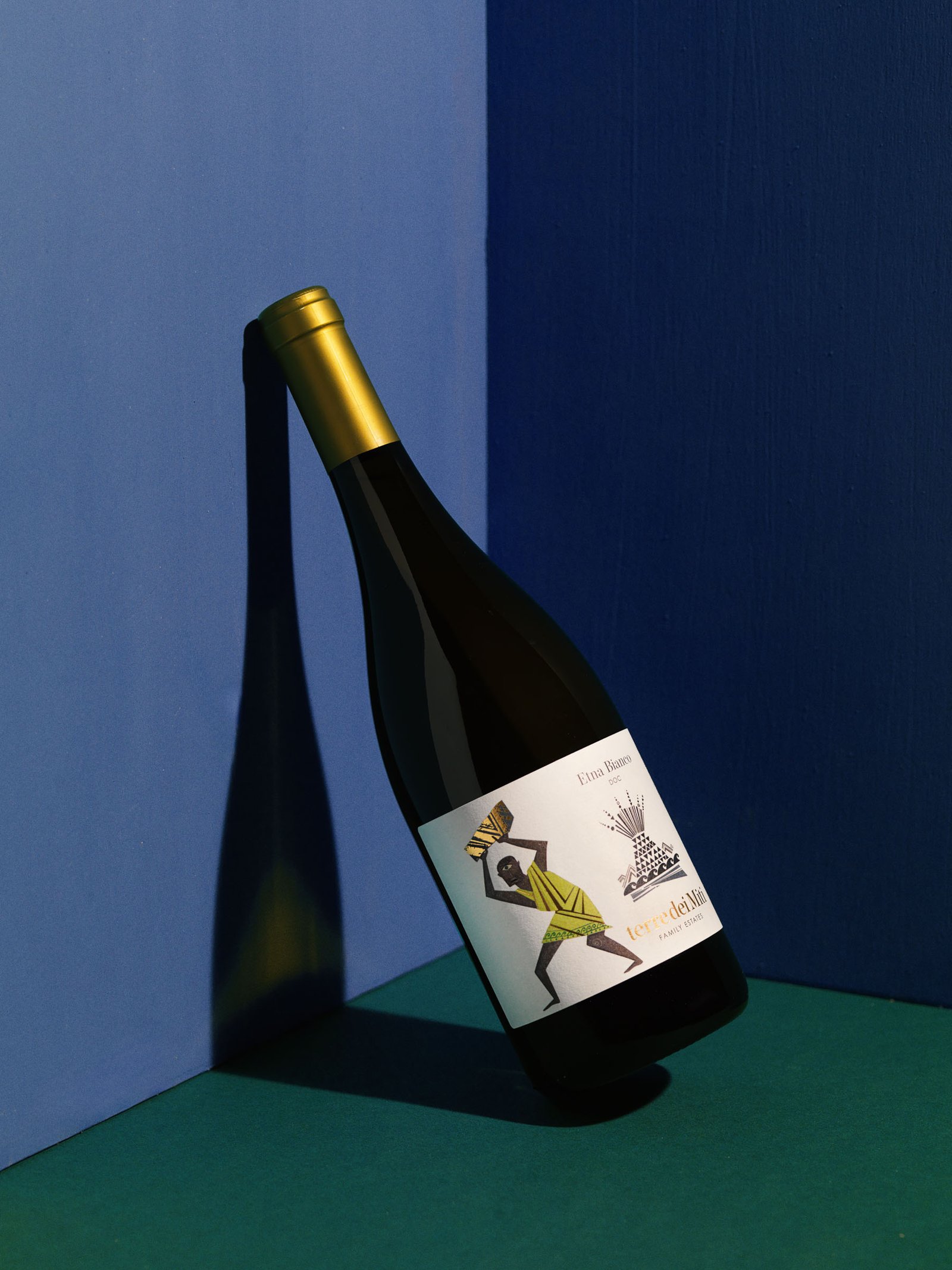 Terre dei Miti Etna Bianco leaning into the corner of two blue walls, on a green surface, with the bottle's long shadow on the wall