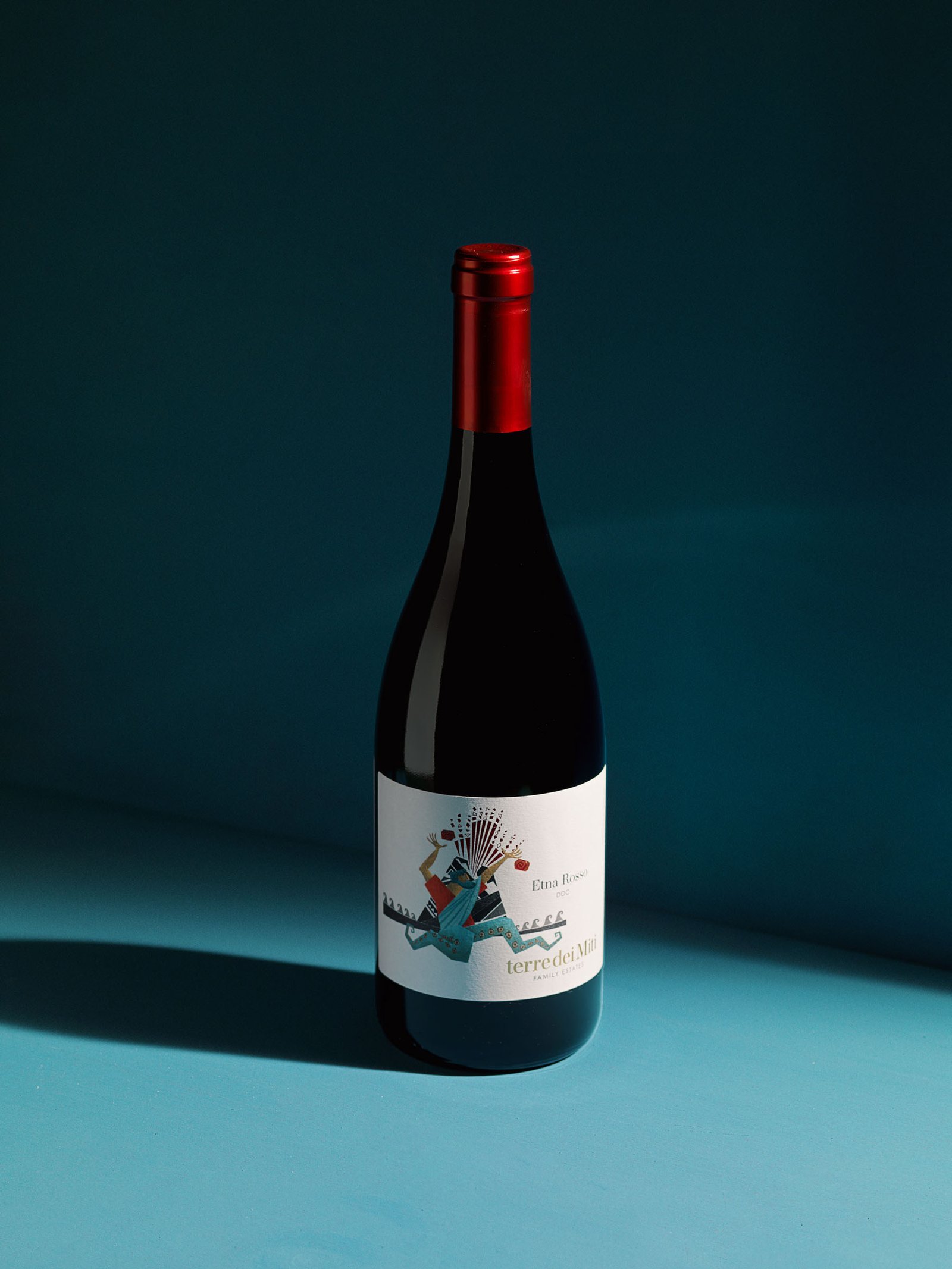 Terre dei Miti Etna Rosso standing on a teal set, in hard light that throws the bottle's shadow