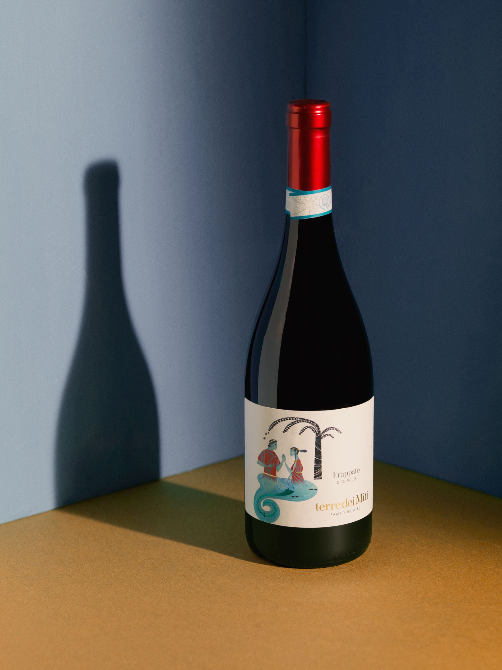 Terre dei Miti Frappato in the corner of two blue walls, on an ochre surface, with the bottle's silhouette in shadow on the wall
