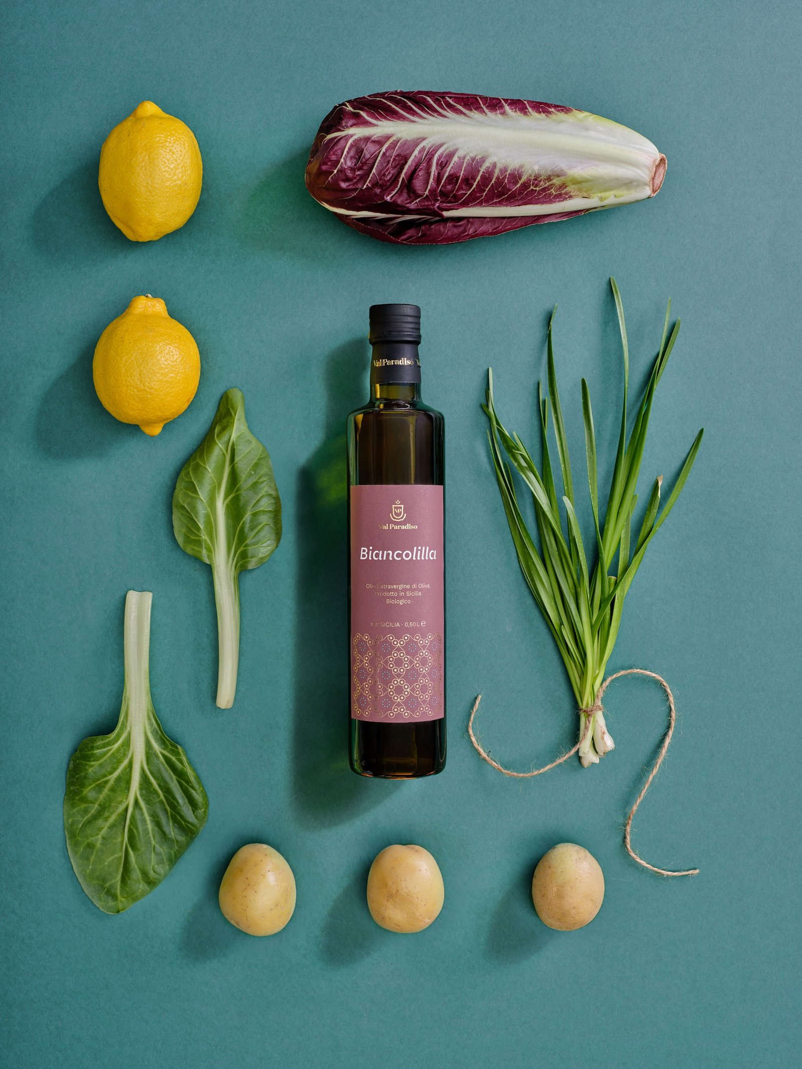 Olio extravergine Val Paradiso Biancolilla, etichetta rosa, coricato su un fondo ottanio fra limoni, radicchio, patate e verdure a foglia disposti in griglia