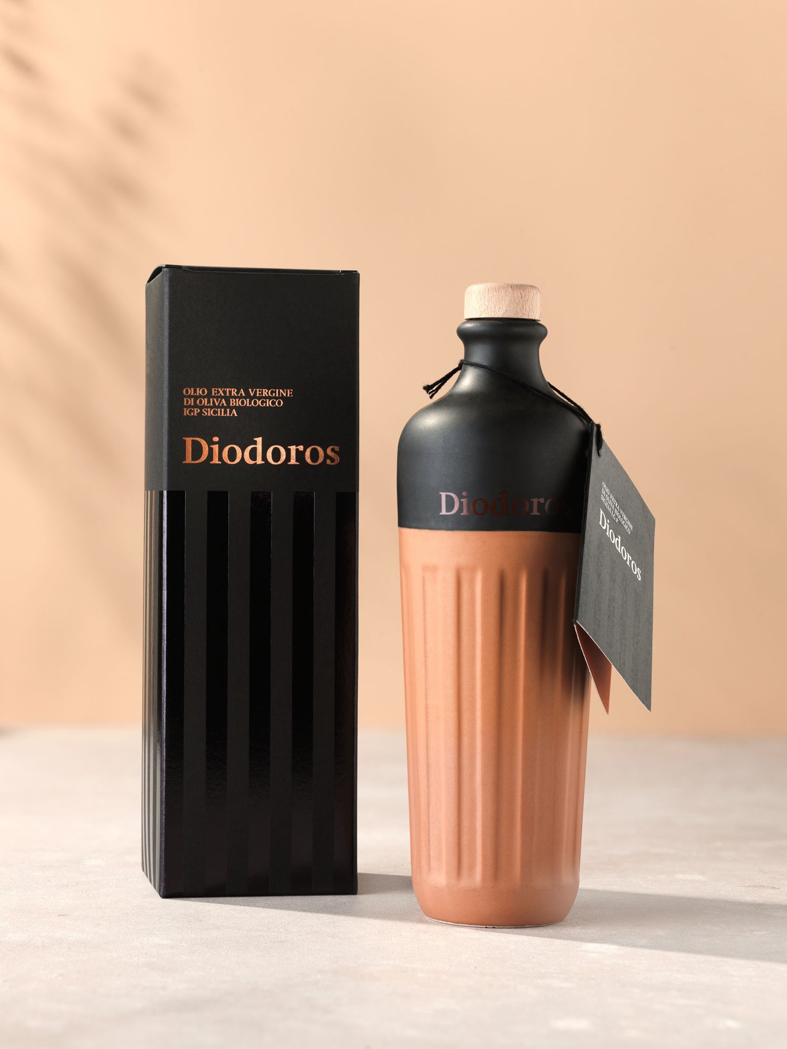 Olio Diodoros di Val Paradiso: la bottiglia di ceramica, nera opaca in alto e color terracotta con le scanalature in basso, accanto all'astuccio nero, sul fondo pesca con l'ombra di un ulivo