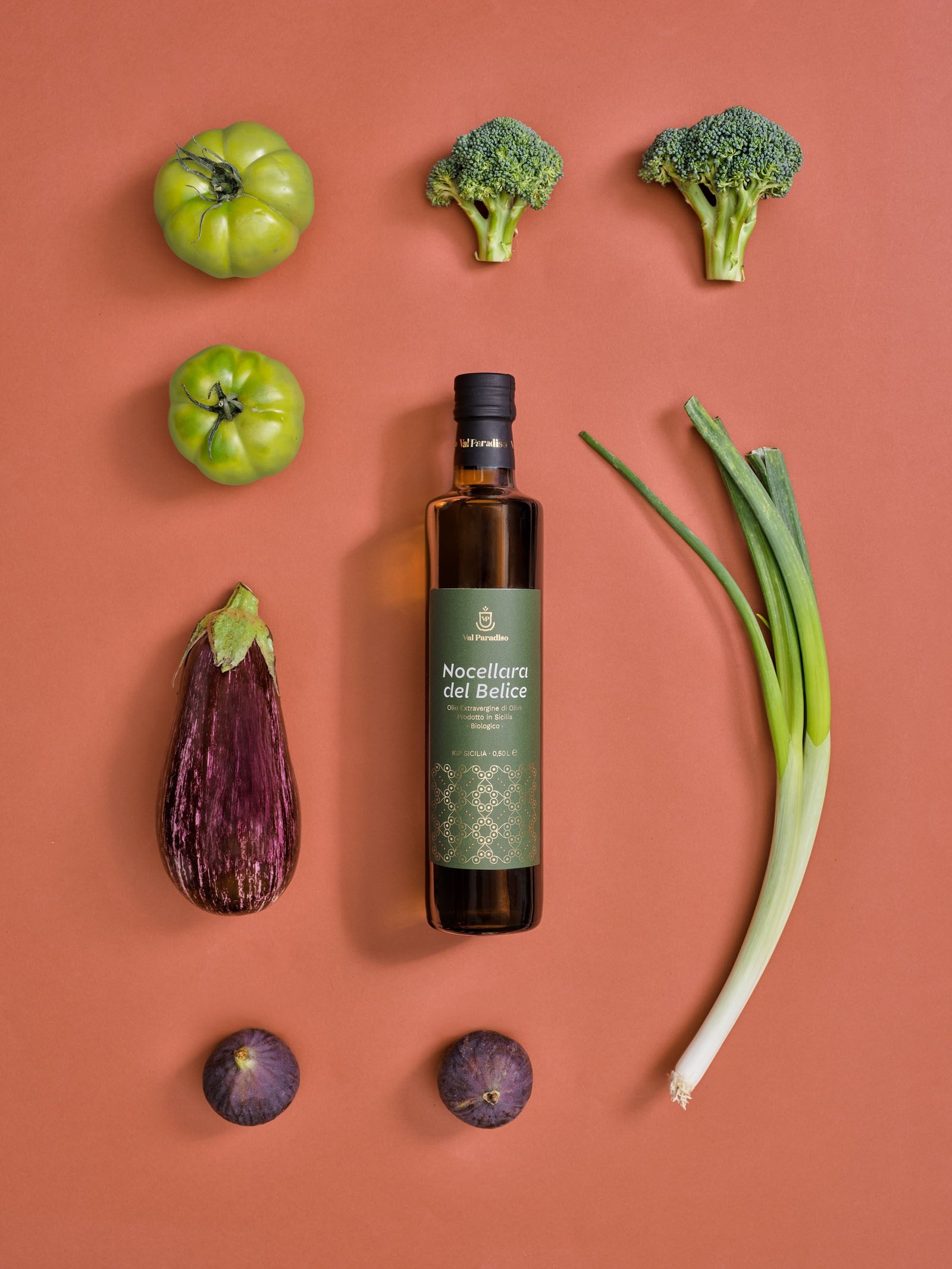 Olio extravergine Val Paradiso Nocellara del Belice, etichetta verde, coricato su un fondo terracotta fra pomodori verdi, broccoli, una melanzana e fichi
