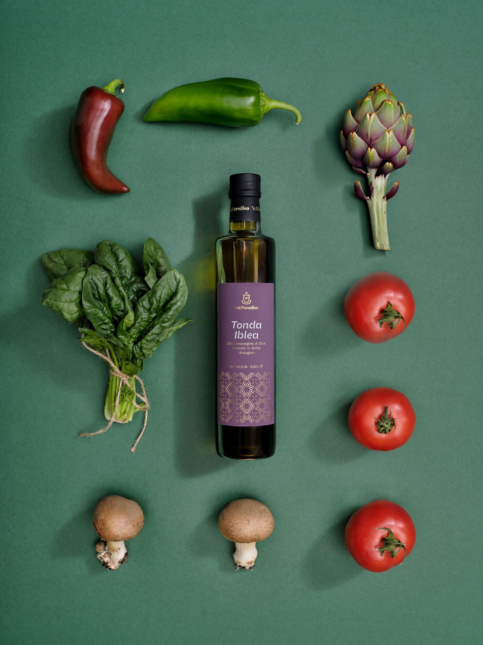 Olio extravergine Val Paradiso Tonda Iblea, etichetta viola, coricato su un fondo verde fra peperoni, un carciofo, spinaci, pomodori e funghi